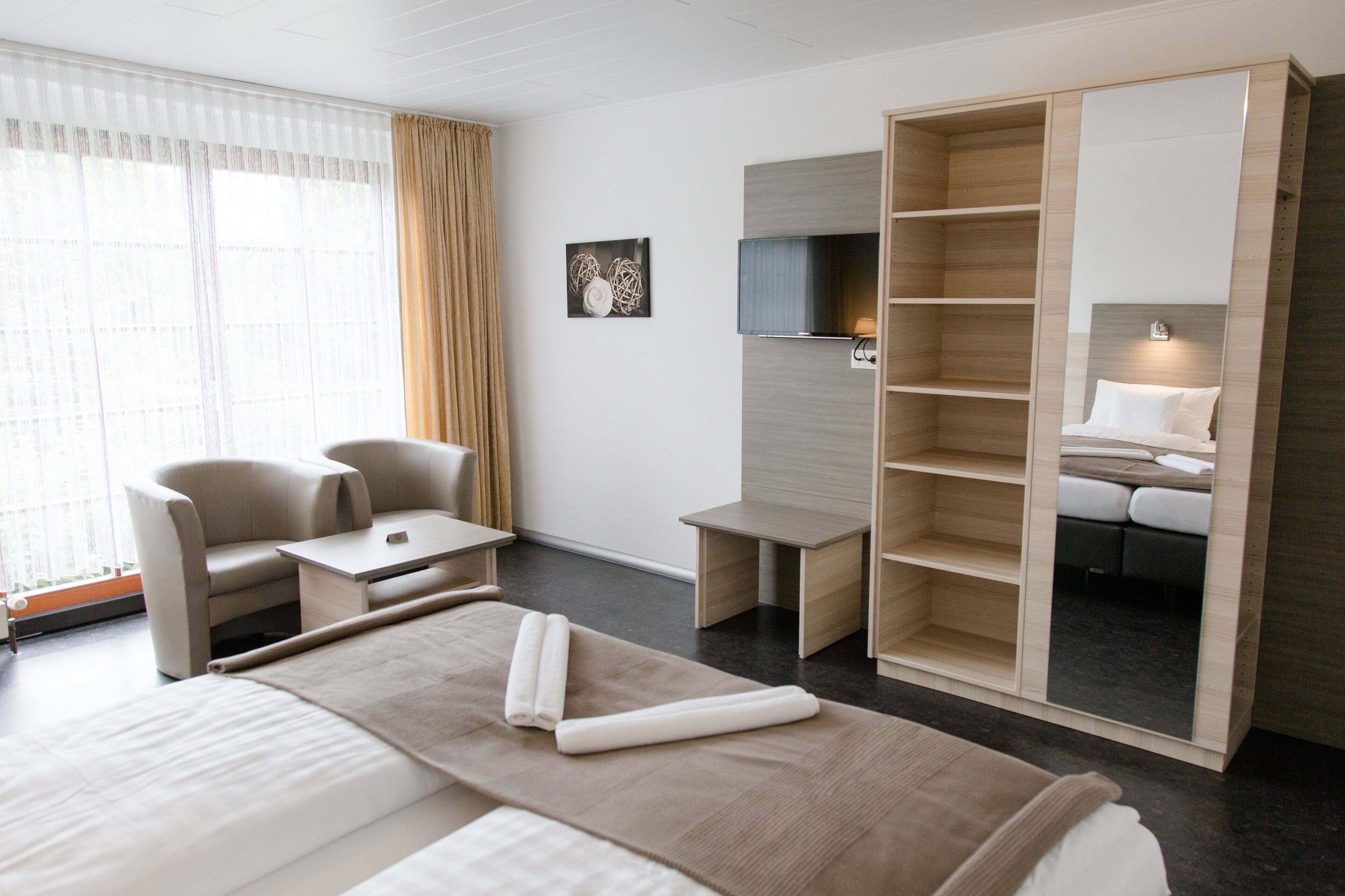 A modern hotel room with a large bed, a seating area with chairs, a TV mounted on the wall, and a wardrobe with a mirror.