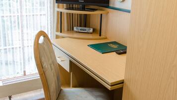 Camera d'albergo con una scrivania in legno, una sedia, una televisione a schermo piatto, una lampada e una finestra con tende.