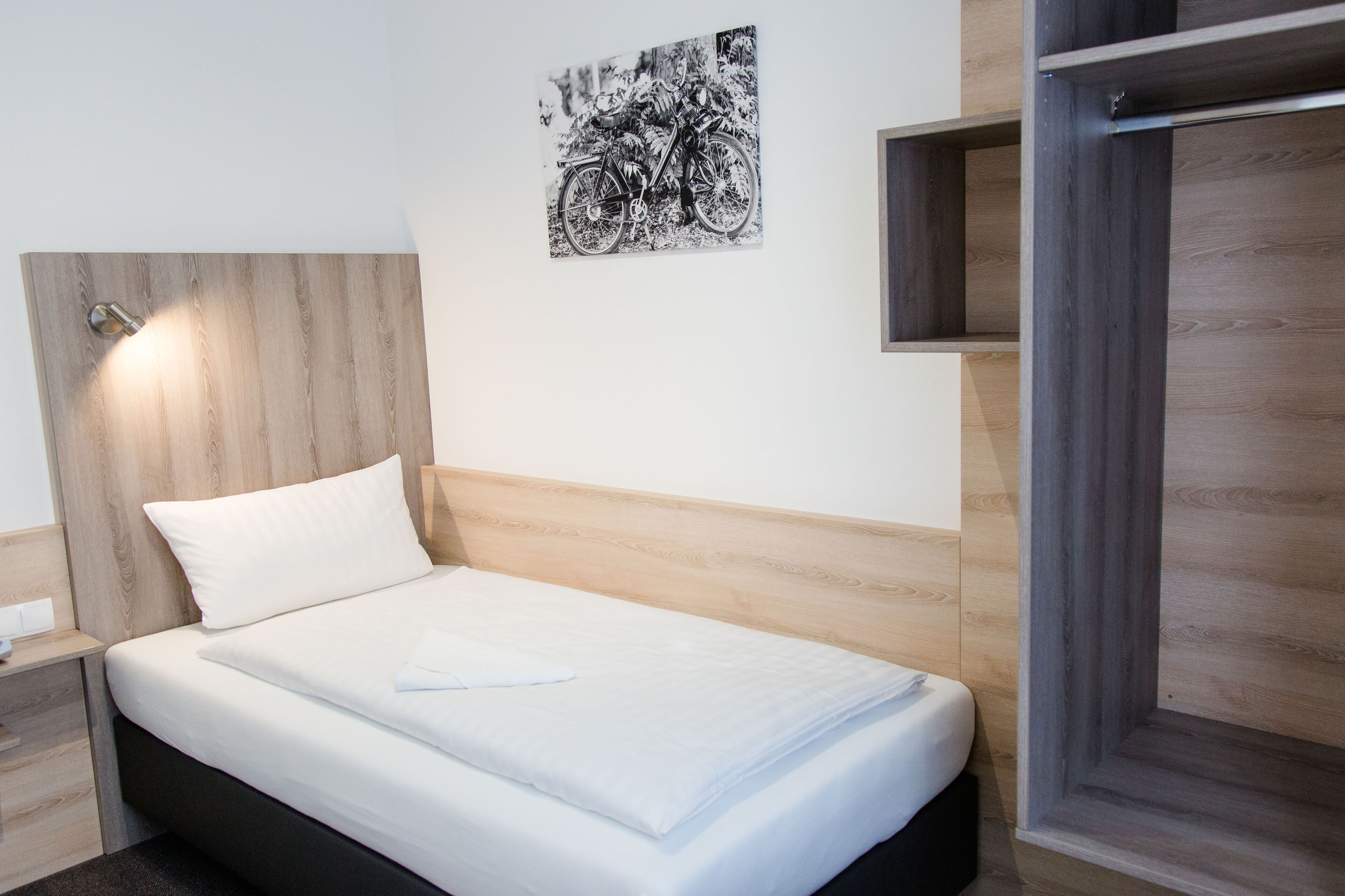 Cama bem arrumada com colchão e travesseiro brancos, cabeceira com acabamento em madeira clara, obra de arte em preto e branco na parede e guarda-roupa de madeira com espelho.