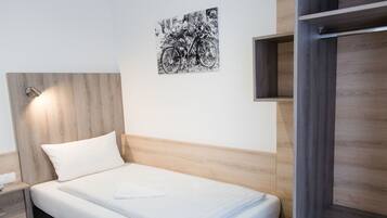 Cama bem arrumada com colchão e travesseiro brancos, cabeceira com acabamento em madeira clara, obra de arte em preto e branco na parede e guarda-roupa de madeira com espelho.