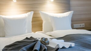 Una cama bien hecha con ropa de cama blanca y gris, una toalla doblada y un objeto decorativo sobre la cama.