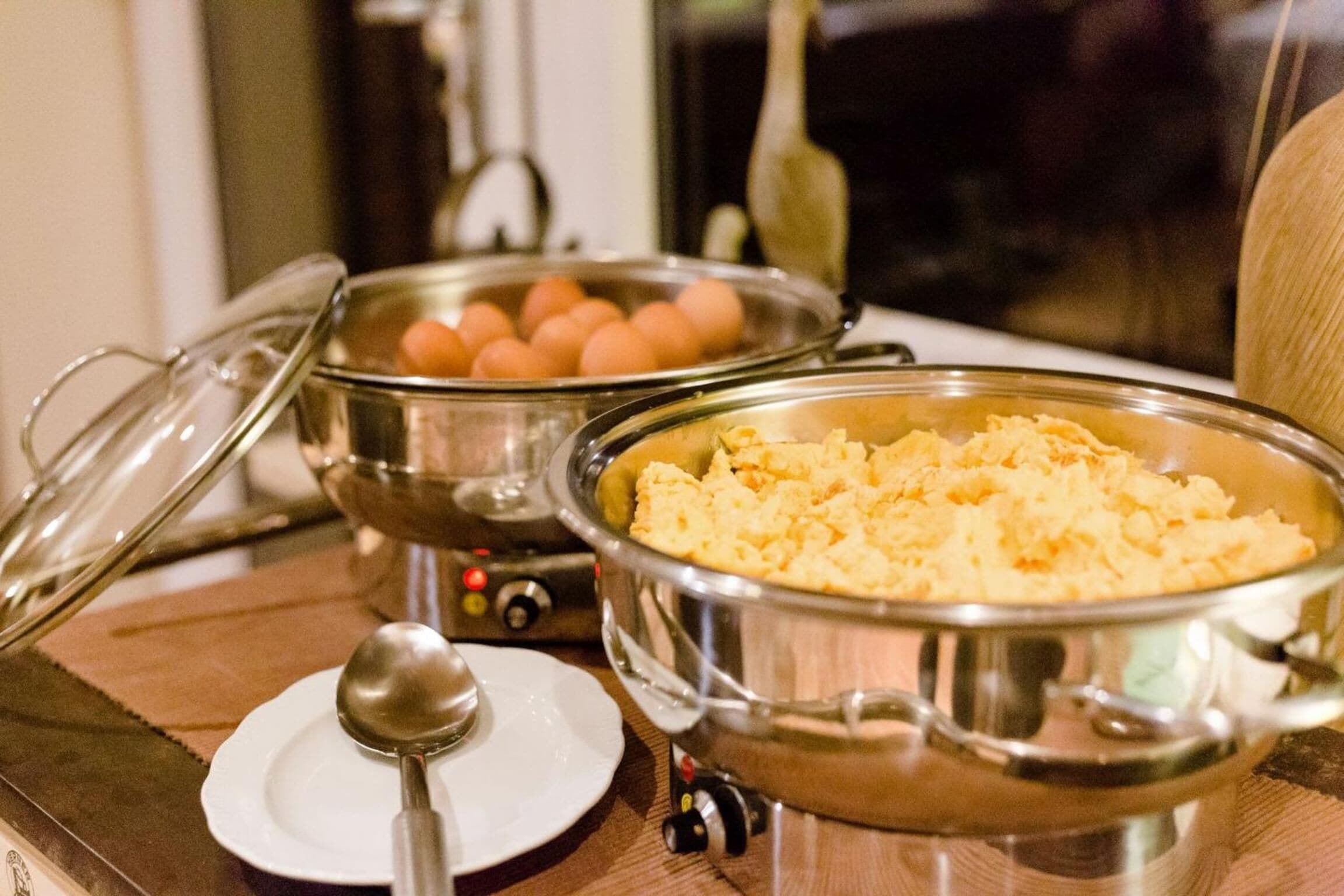 A buffet setup with various dishes in metal containers, a spoon on a plate, and a window in the background.
