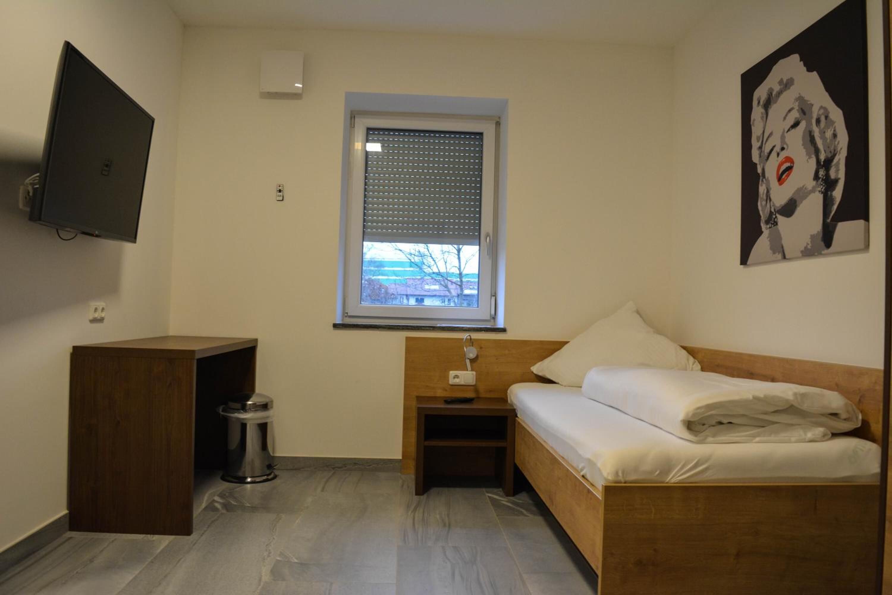 A small, neatly arranged bedroom with a single bed, a bedside table, a TV mounted on the wall, a window with blinds, and a poster on the wall.