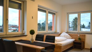 Een moderne kamer met een houten bureau, twee zwarte stoelen, een bed met witte beddengoed en een klein nachtkastje met een plant.