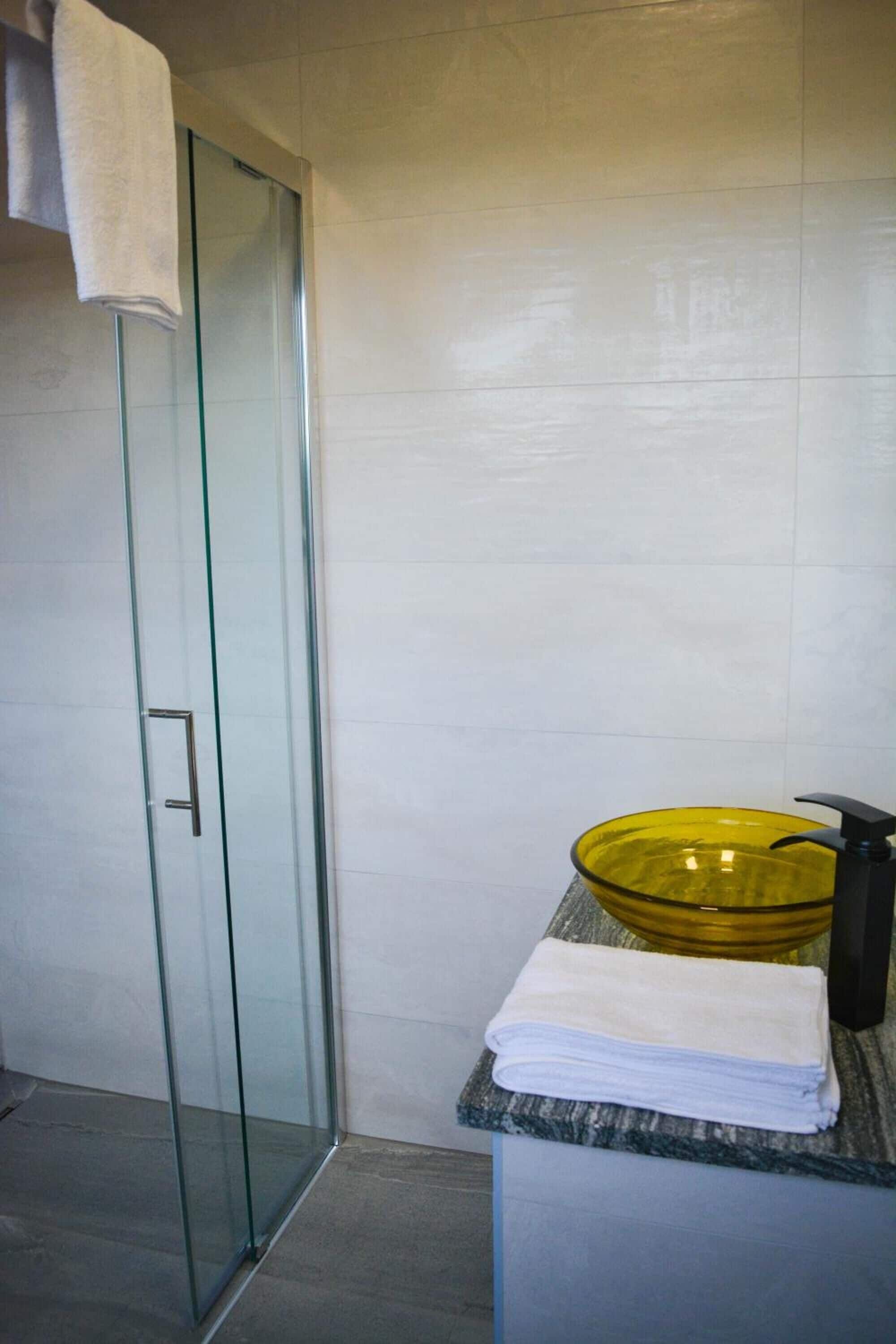 A bathroom with a glass shower door, a yellow glass bowl sink, and a white towel on a marble countertop.