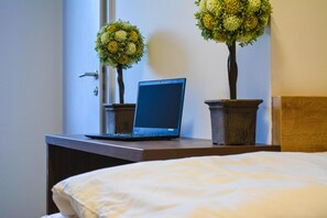 Una laptop sobre un escritorio de madera con dos plantas en macetas, una cama con cubrecama blanco y un cabecero de madera.