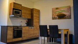 Een kleine keuken met houten kastjes, een magnetron en een oven. Er staat een eettafel met stoelen en er hangt een schilderij aan de muur.