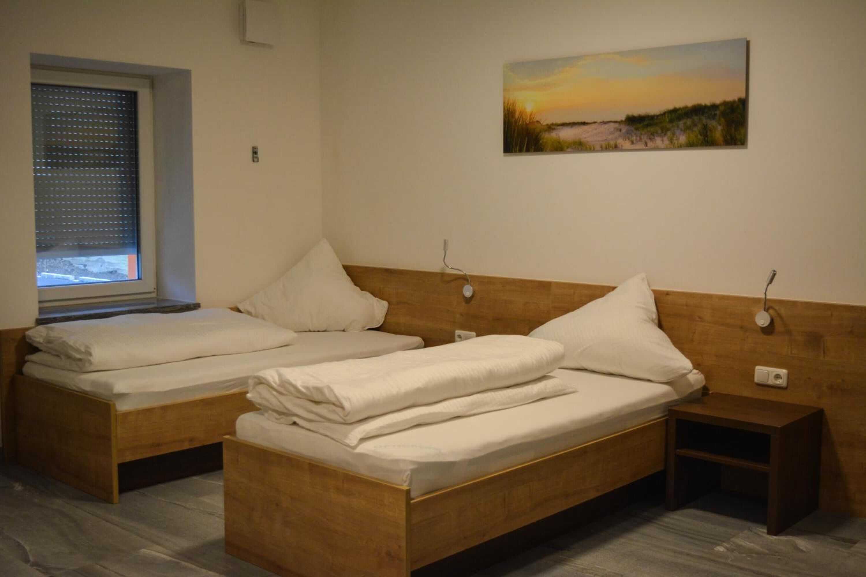Una habitación con dos camas individuales, una mesita de noche, un velador, una ventana con persianas y un cuadro en la pared.