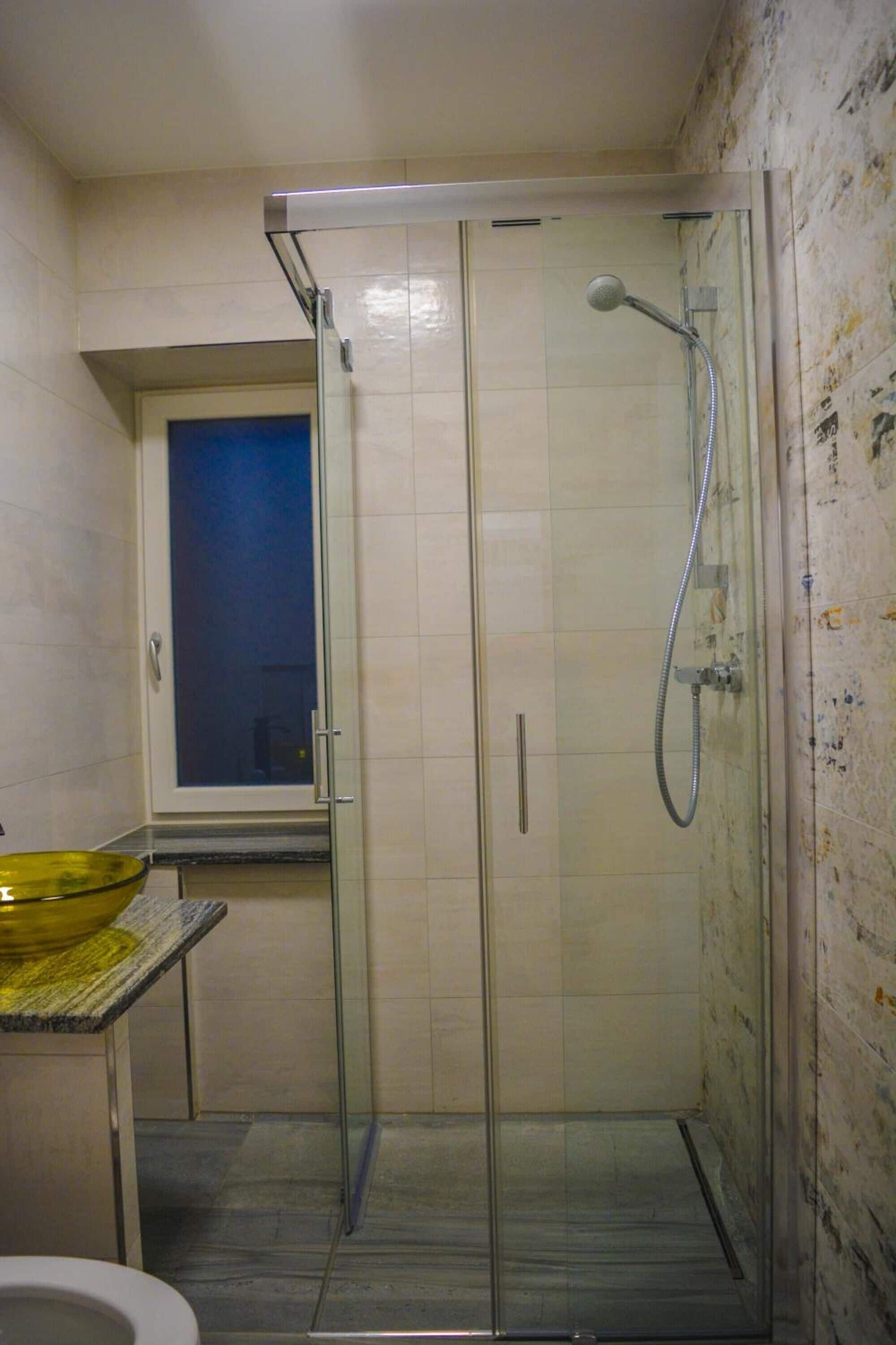 Un área de ducha pequeña y cerrada con puerta de cristal, ventana y lavamanos.