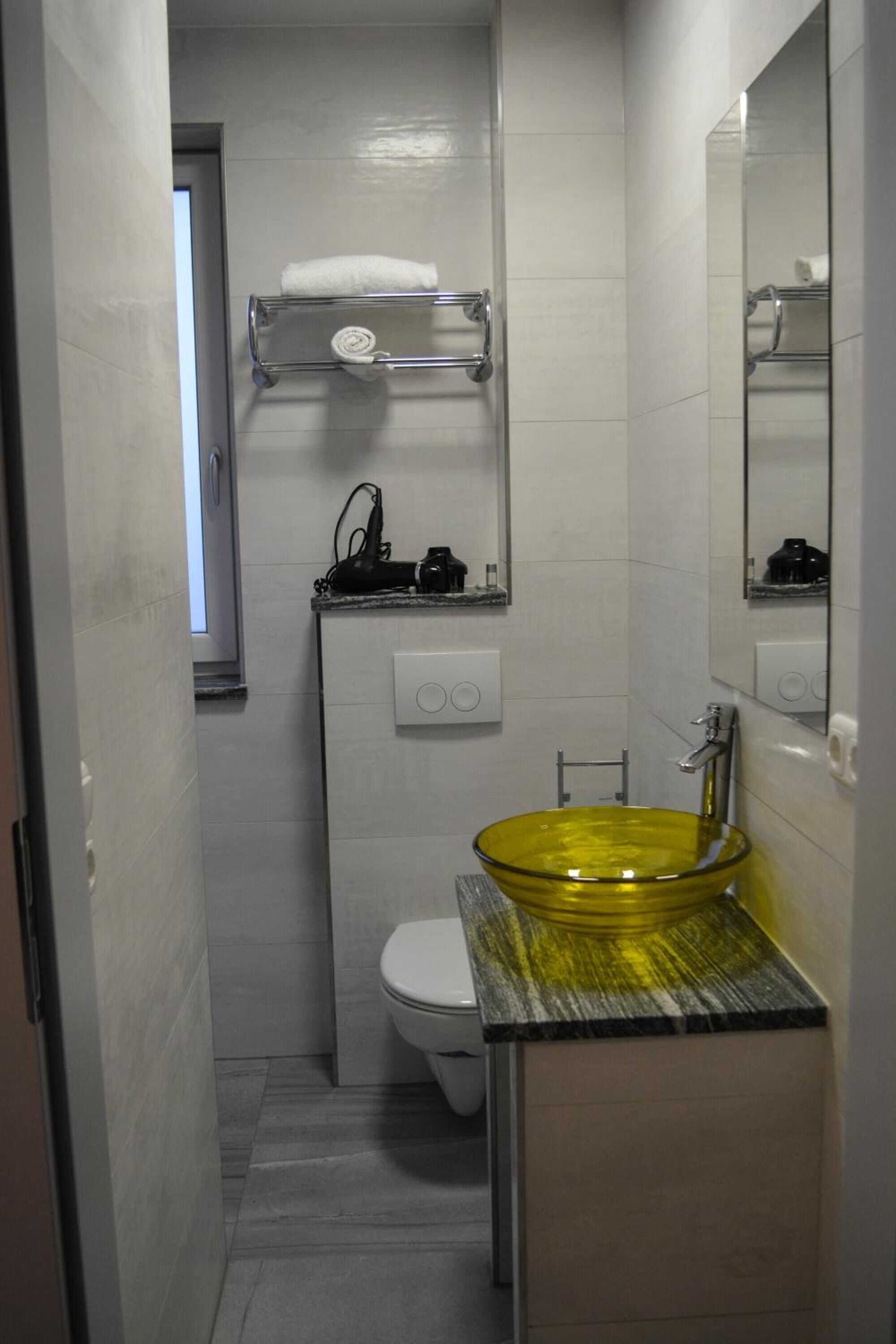 Un baño pequeño con un lavamanos de vidrio amarillo, un inodoro blanco y un perchero sobre el inodoro.