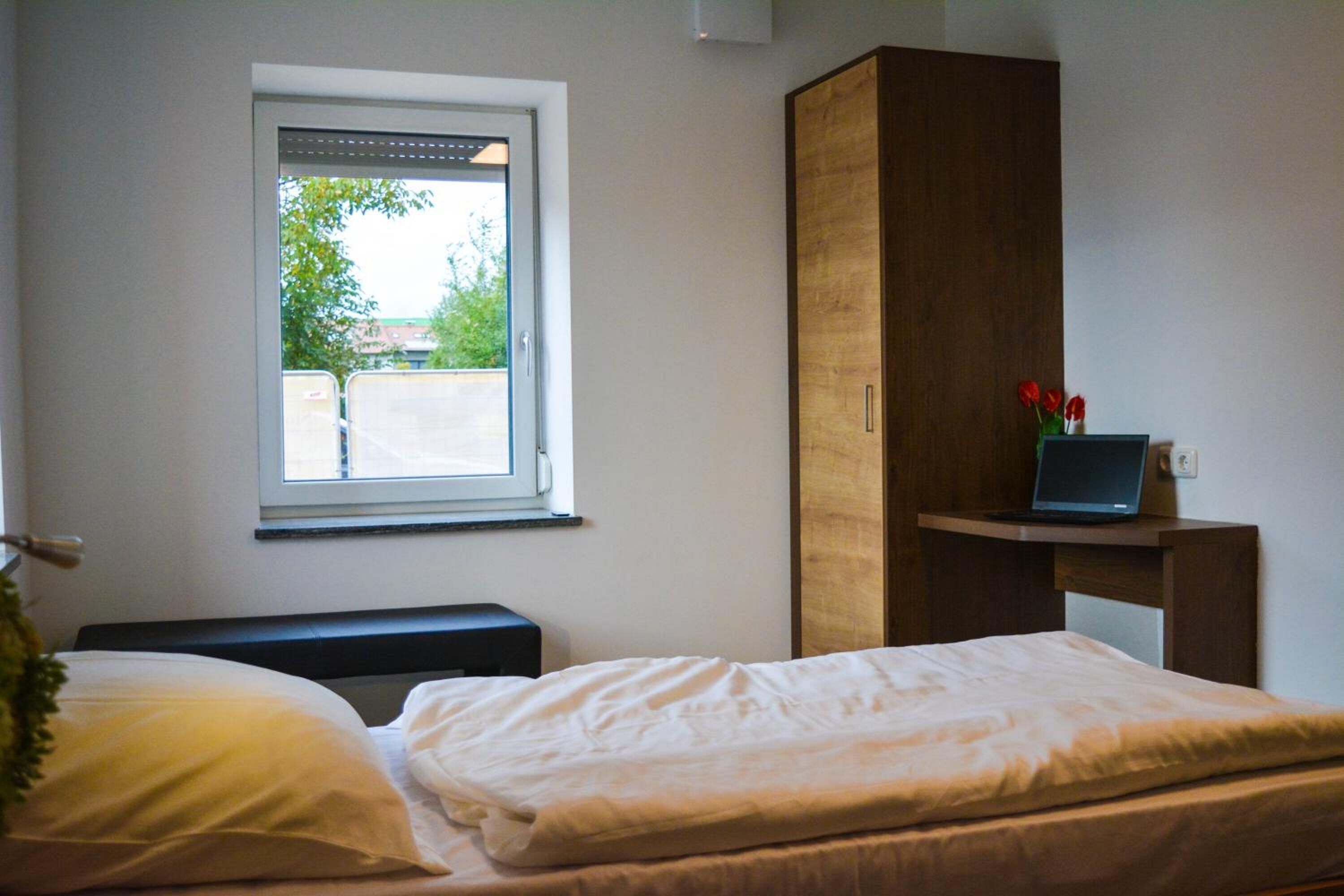 A bedroom with a bed, a window, a wooden wardrobe, and a laptop on a small table.