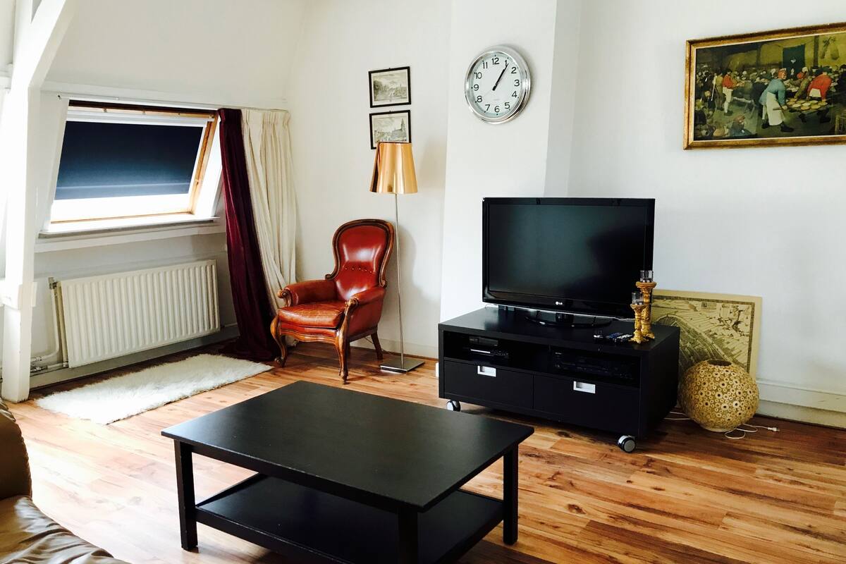 A living room with a wooden floor, a black TV stand, a leather armchair, a black coffee table, and a window with curtains.
