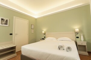 Habitación de hotel ordenada con cama, lámparas de noche, mesita de noche, puerta y estante fijado a la pared.