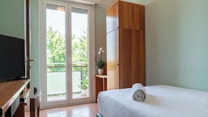 A hotel room with a bed, a television, a wooden wardrobe, a potted plant, and a sliding glass door leading to an outdoor area.