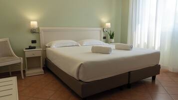 A neatly made bed with white linens and two pillows, a bedside table with a lamp, a chair, and a potted plant.