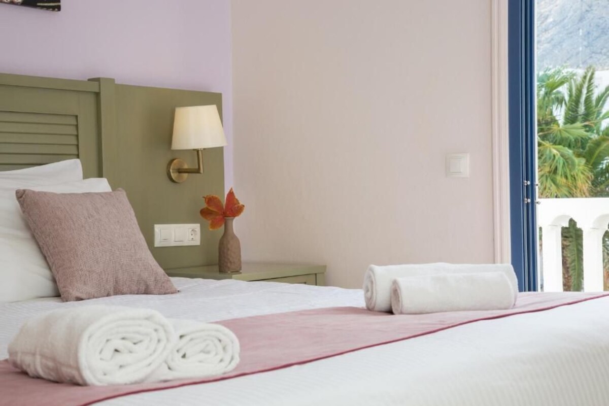 A hotel room with a bed, a nightstand with a lamp, a vase with flowers, and a window with a view of palm trees.