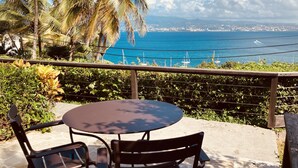 Eine Terrasse mit rundem Tisch und zwei Stühlen, mit Blick auf eine Küstenlandschaft mit Booten und einer entfernten Stadtansicht.