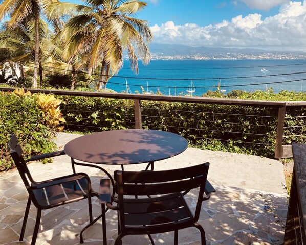A patio with a round table and two chairs, overlooking a coastal view with boats and a distant cityscape. - Habitation Desrosiers (Trois-Ilets)