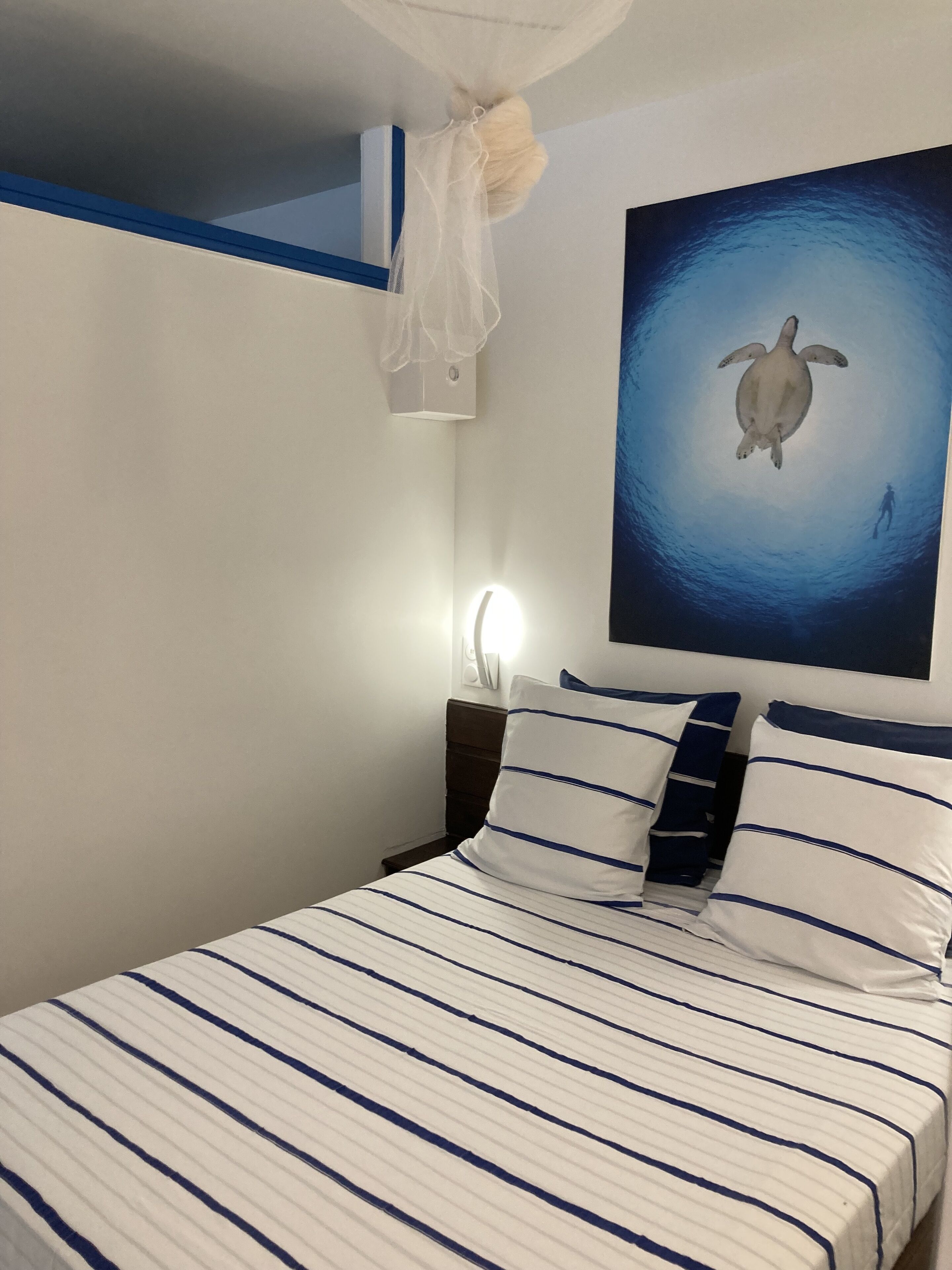 A bedroom with a bed featuring striped bedding, a wall-mounted light, and a painting of a turtle.