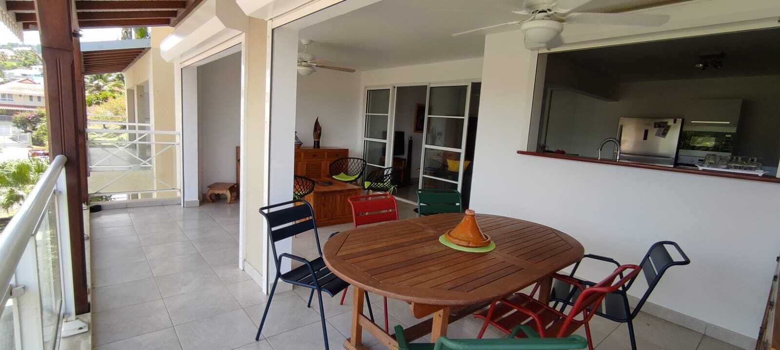 A covered outdoor dining area with a wooden table and chairs, a ceiling fan, and a view of the surrounding buildings.