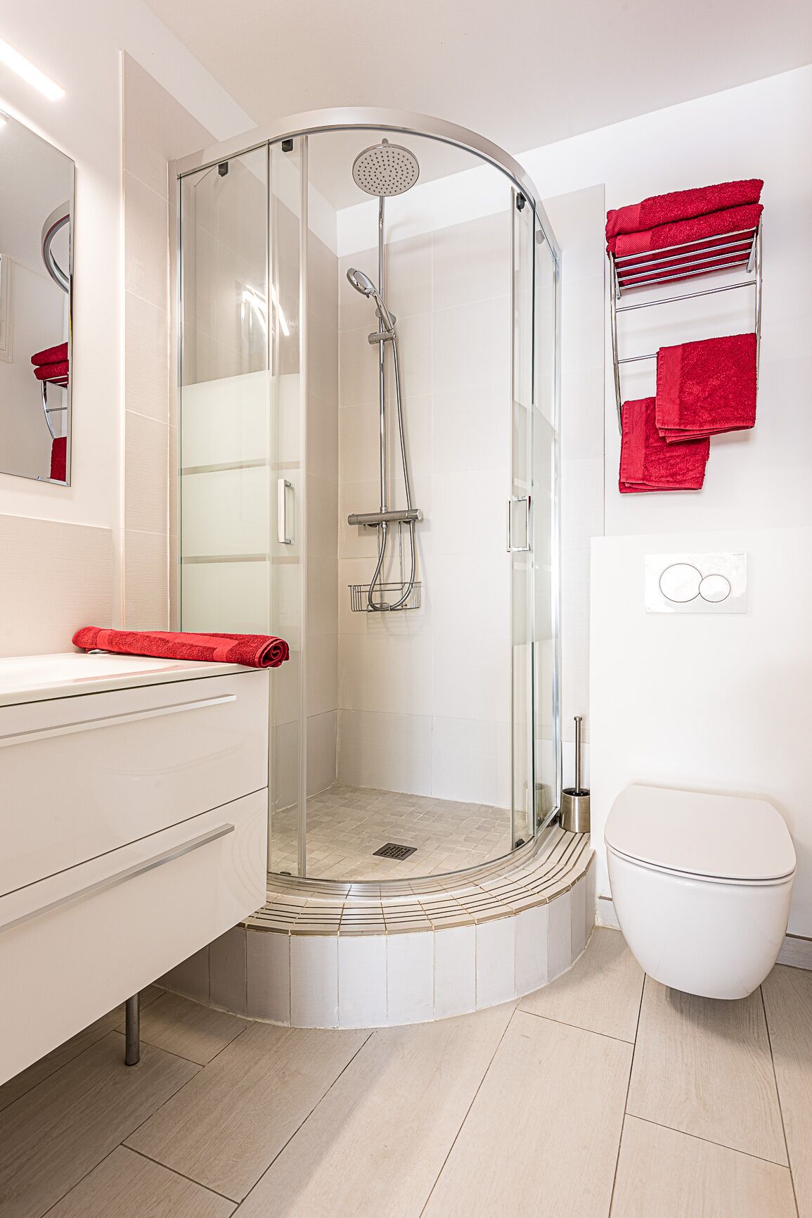 Un baño moderno con ducha envuelta en vidrio, inodoro blanco y tocador con una toalla roja.