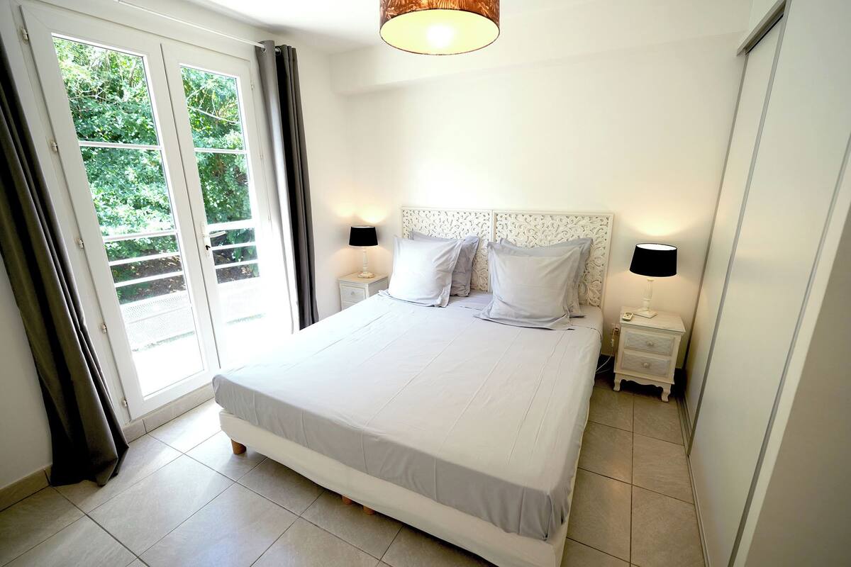 A bedroom with a bed, two bedside tables with lamps, a window with curtains, and a view of greenery outside.