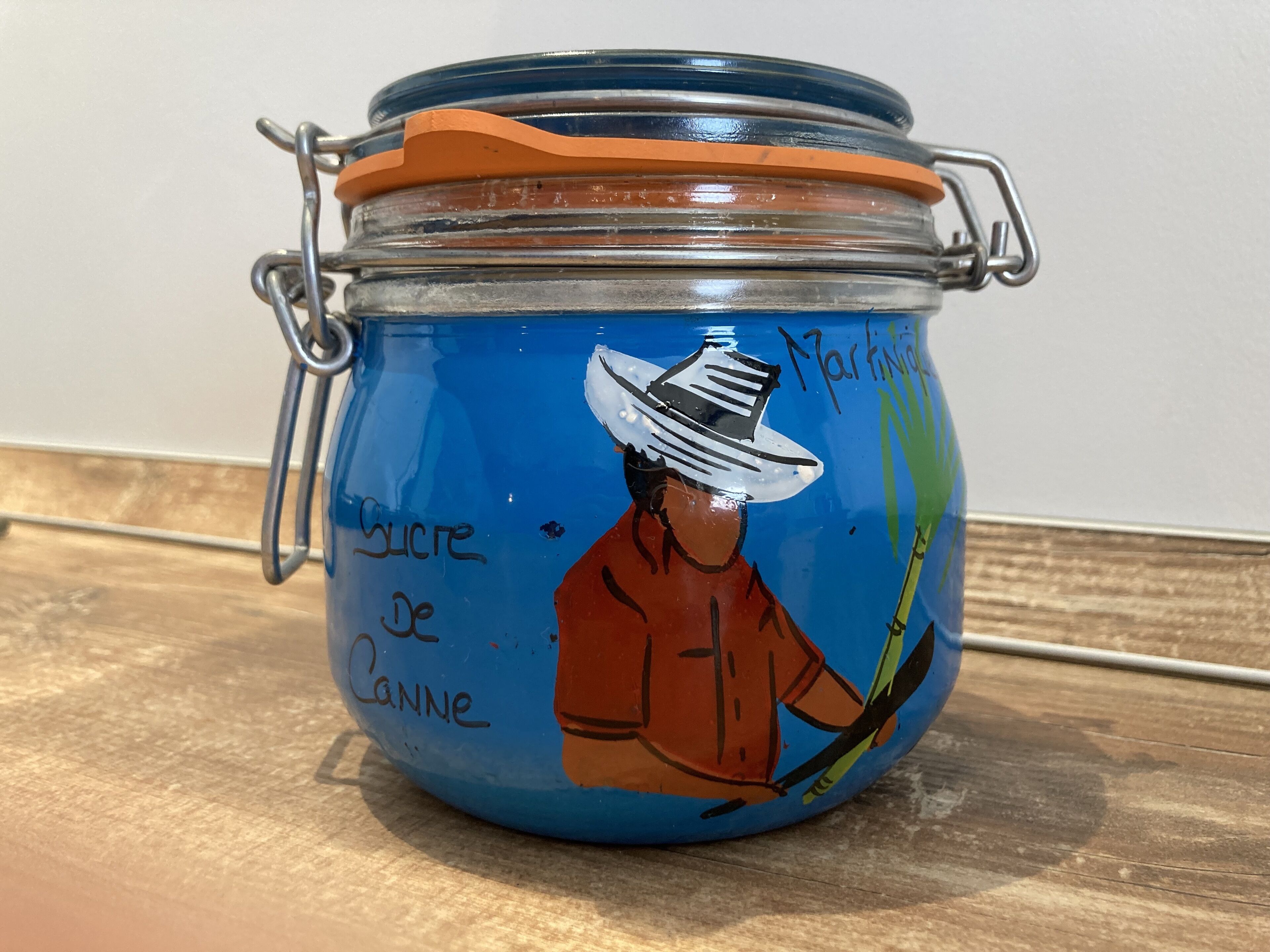 A glass jar with a metal clasp lid, featuring a drawing of a person wearing a hat and holding a cane, with the text "Sucre de Canne" and "Martinique" visible.
