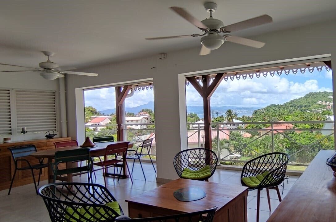 A spacious room with a dining area, a ceiling fan, large windows with a view of a hilly landscape.