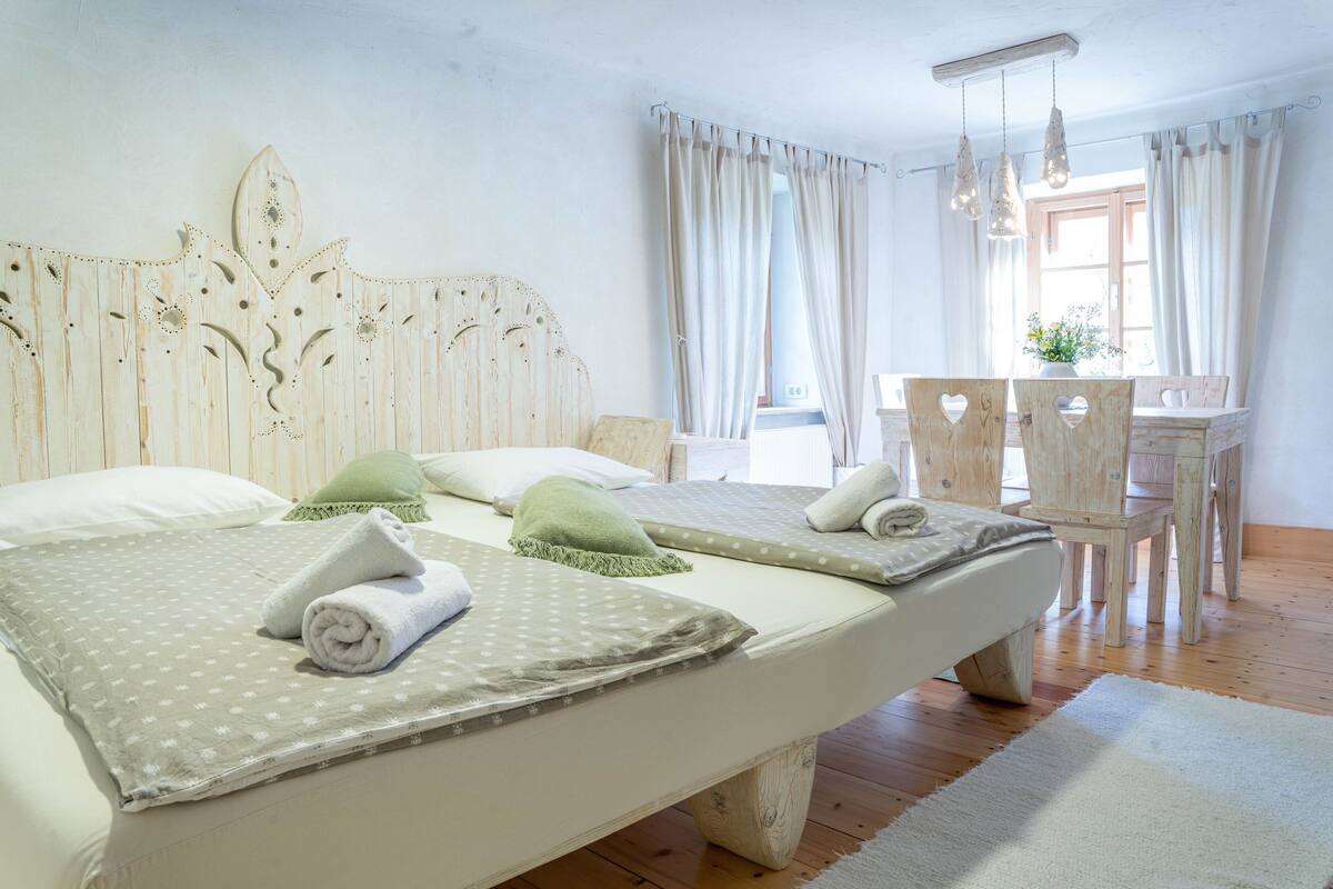 Ein Doppelbett mit Holz-Kopfteil, zwei Kissen und einer grünen Tagesdecke. Ein Holzstuhl und ein kleiner Tisch mit einer Pflanze stehen daneben.