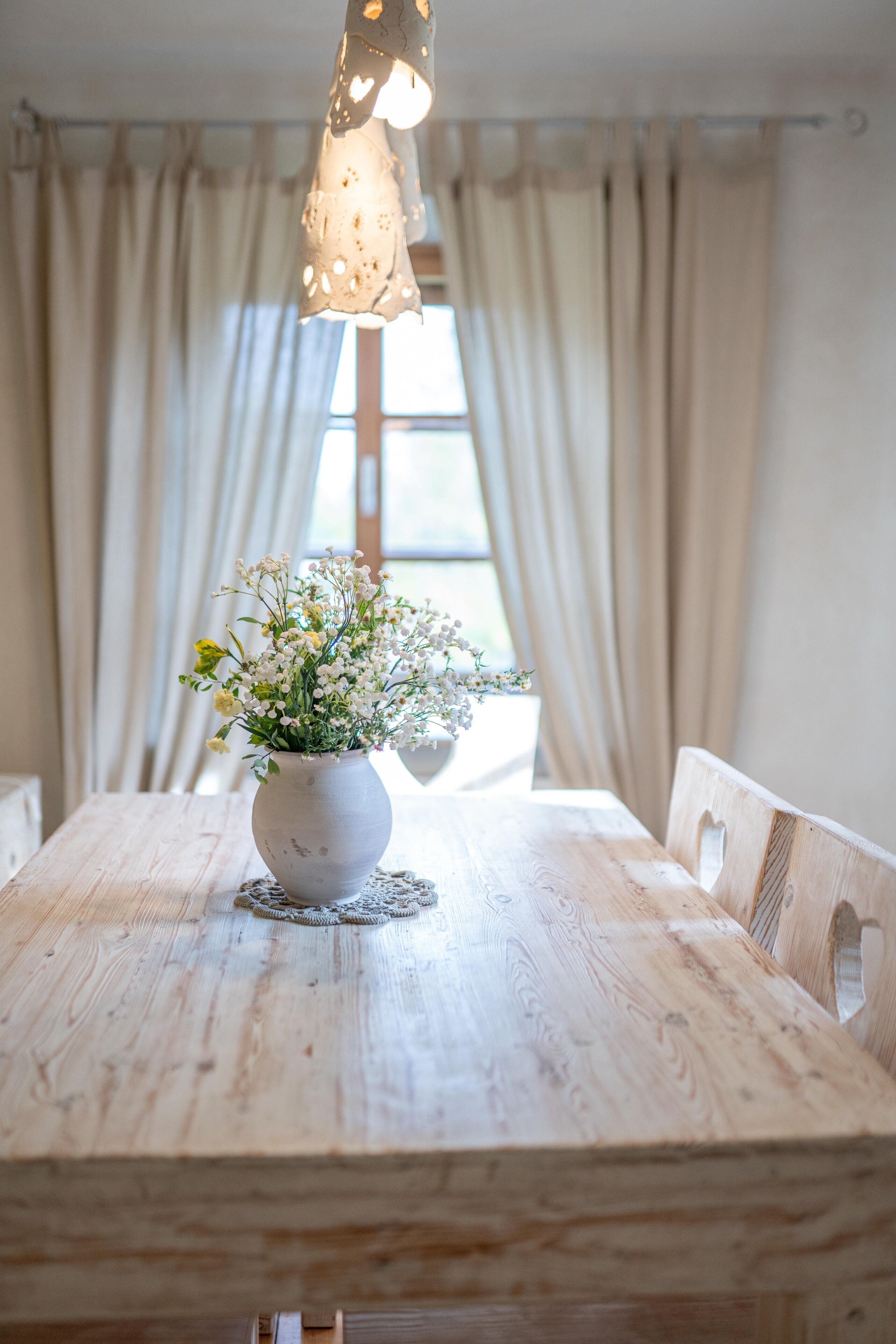 A wooden dining table with a white vase of flowers, a woven placemat, and a hanging light fixture.