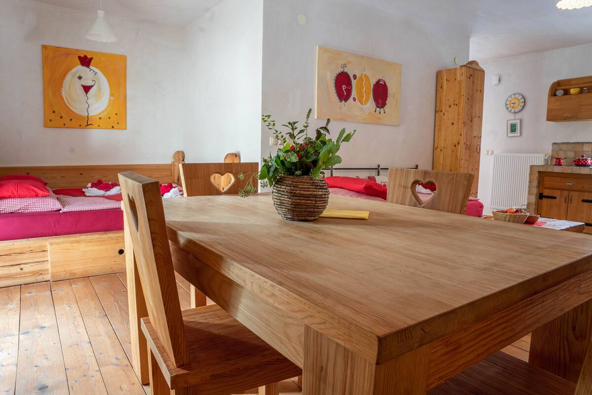 Ein Holzesstisch mit Stühlen, eine Vase mit Blumen und ein rotkariertes Sofa in einem Raum mit Holzwänden und einem Küchenbereich im Hintergrund.