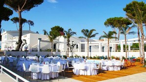An outdoor dining area with round tables and chairs, set against a backdrop of a white building and palm trees. - Agriturismo Campo Allegro (Santa Ninfa)