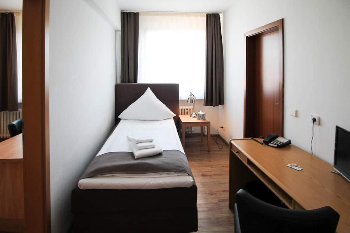 A hotel room with a single bed, a desk with a computer, a chair, and a telephone.