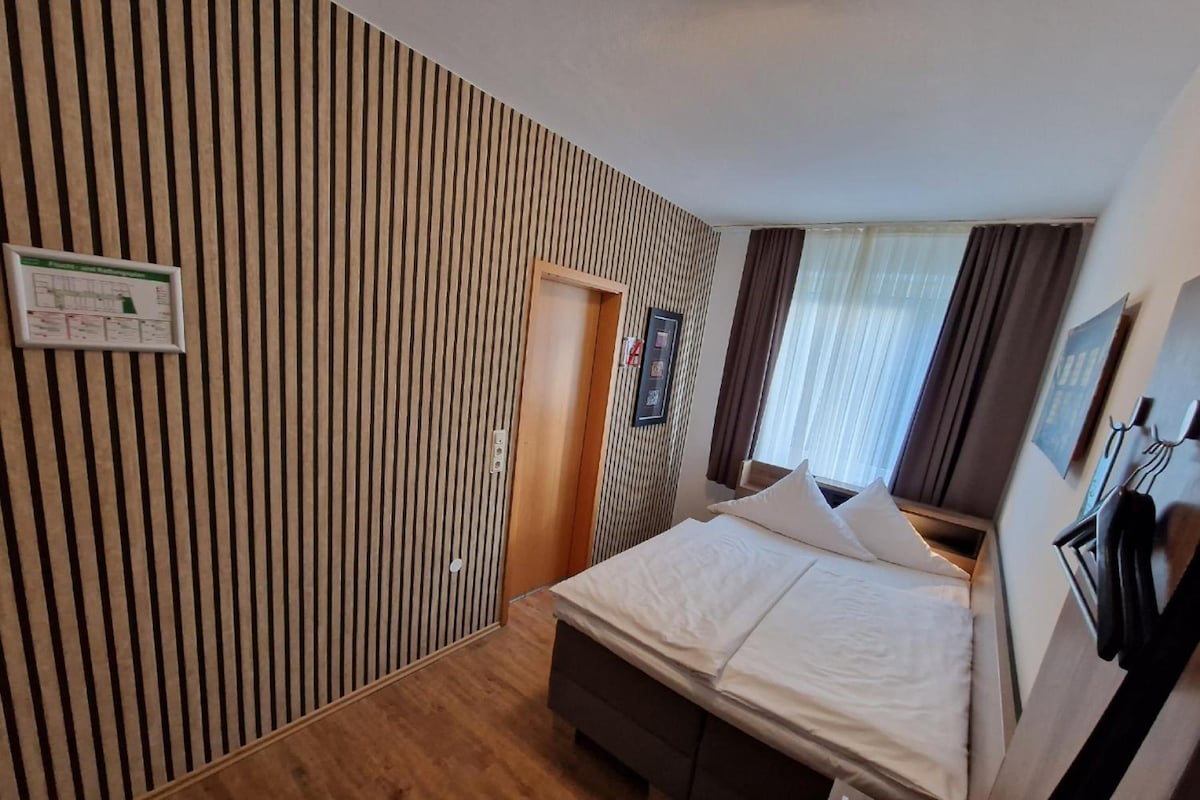 Une chambre d’hôtel avec un plancher en bois, un lit avec du linge de maison blanc, une porte en bois et une fenêtre avec des rideaux.
