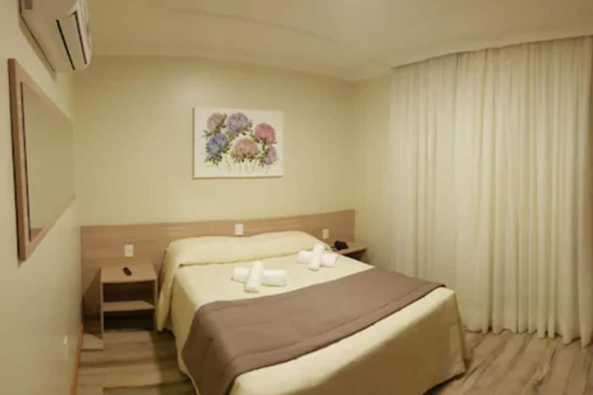 A hotel room with a bed, a nightstand, a painting of flowers, and a window with curtains.