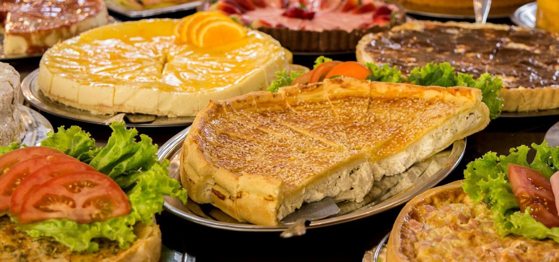 A variety of pies and desserts on a table, surrounded by fresh vegetables like lettuce and tomatoes.