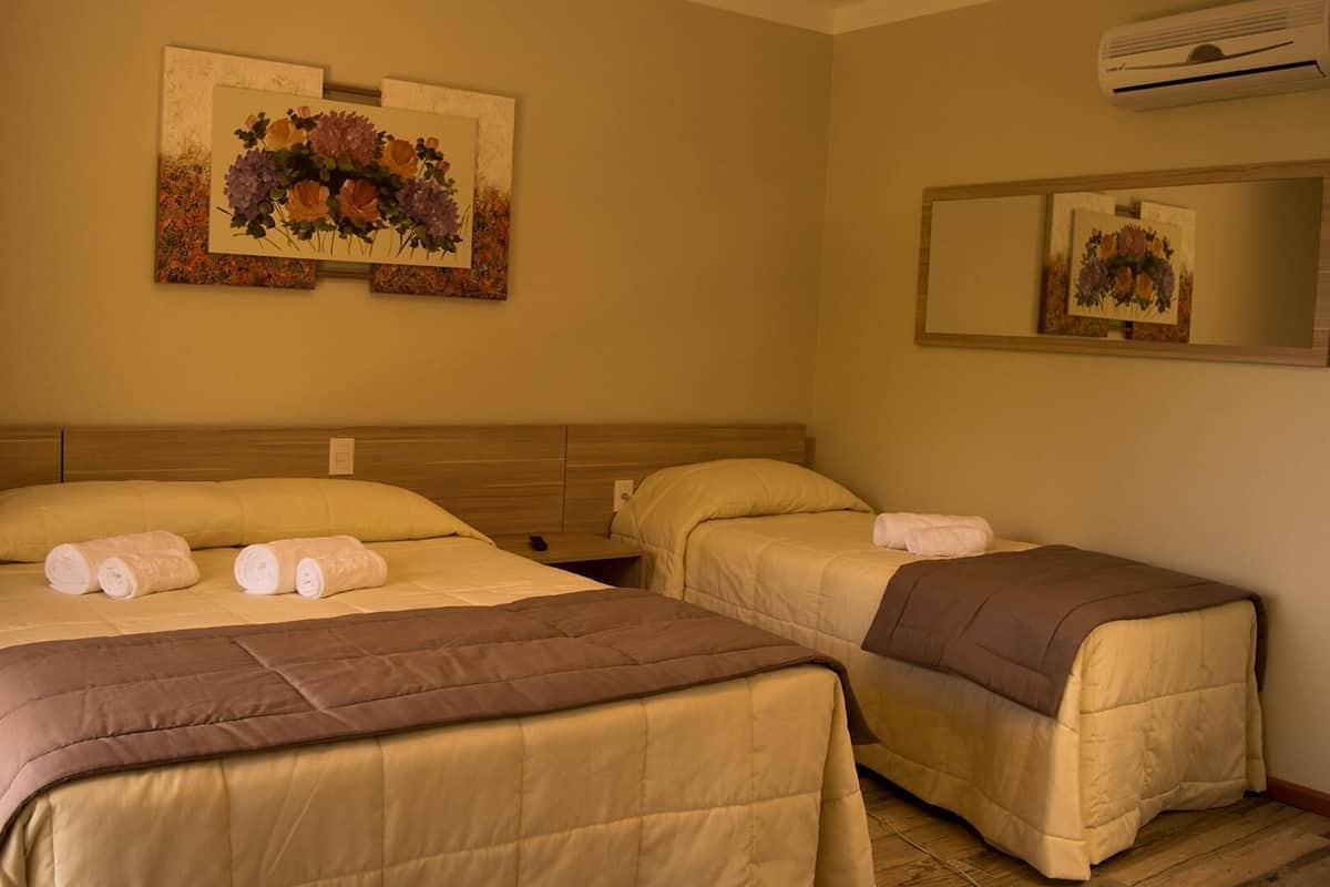 A hotel room with two beds, beige bedding, a wooden headboard, a wall-mounted air conditioner, and a floral painting.