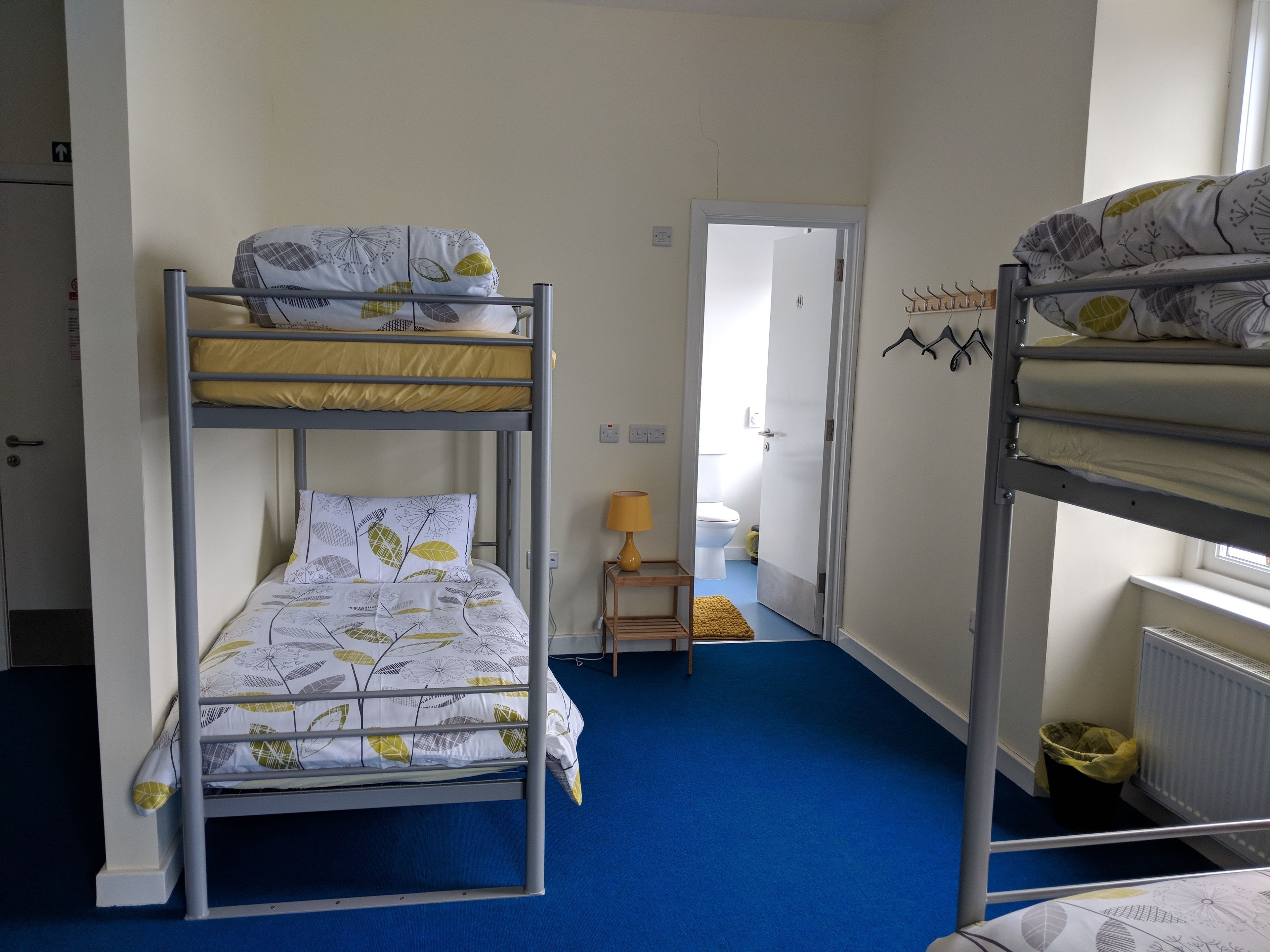 A bunk bed room with a blue carpet, a small bedside table with a lamp, and a door leading to a bathroom.