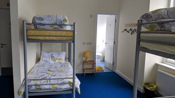 A bunk bed room with a blue carpet, a small bedside table with a lamp, and a door leading to a bathroom.