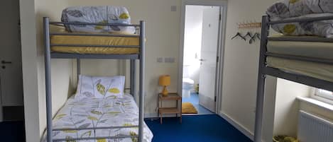 Habitación con literas, una alfombra azul, una mesita de noche con lámpara y una puerta que da al baño.