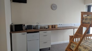Een kleine keuken met een magnetron, broodrooster, waterkoker en opbergcontainers. Een klok hangt aan de muur, en een houten stoel staat bij het raam.