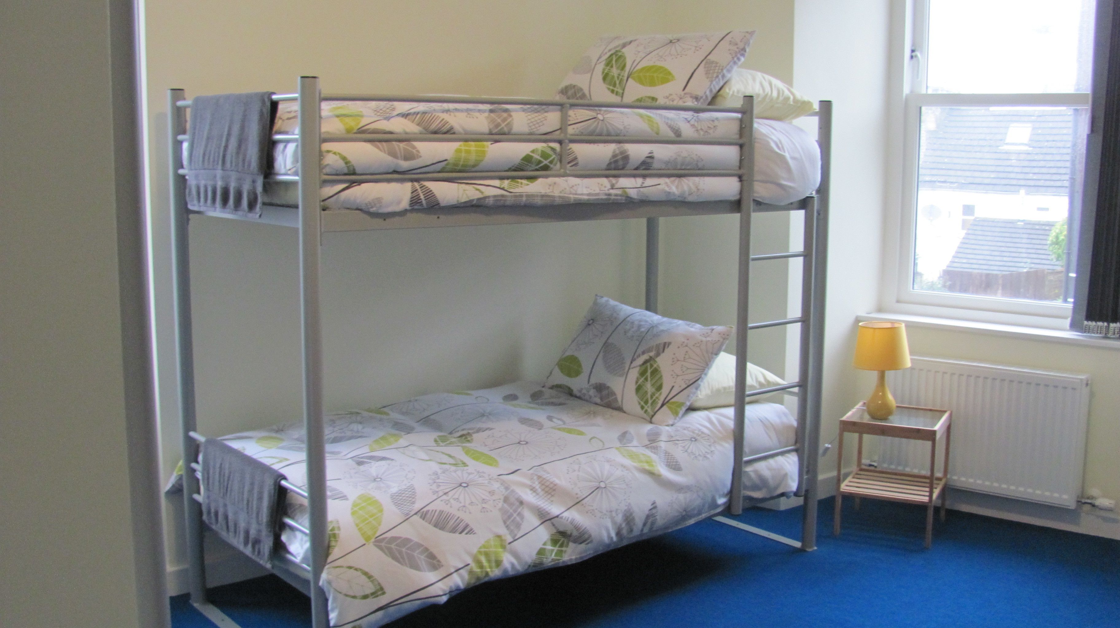Een stapelbed met beddengoed met bladmotieven, een klein nachtkastje met een lamp en een raam met uitzicht op gebouwen.