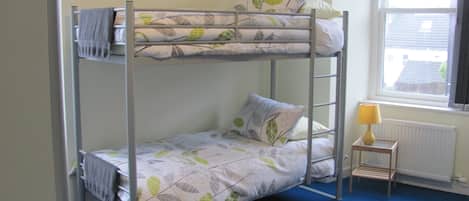 A bunk bed with leaf-patterned bedding, a small bedside table with a lamp, and a window with a view of buildings.