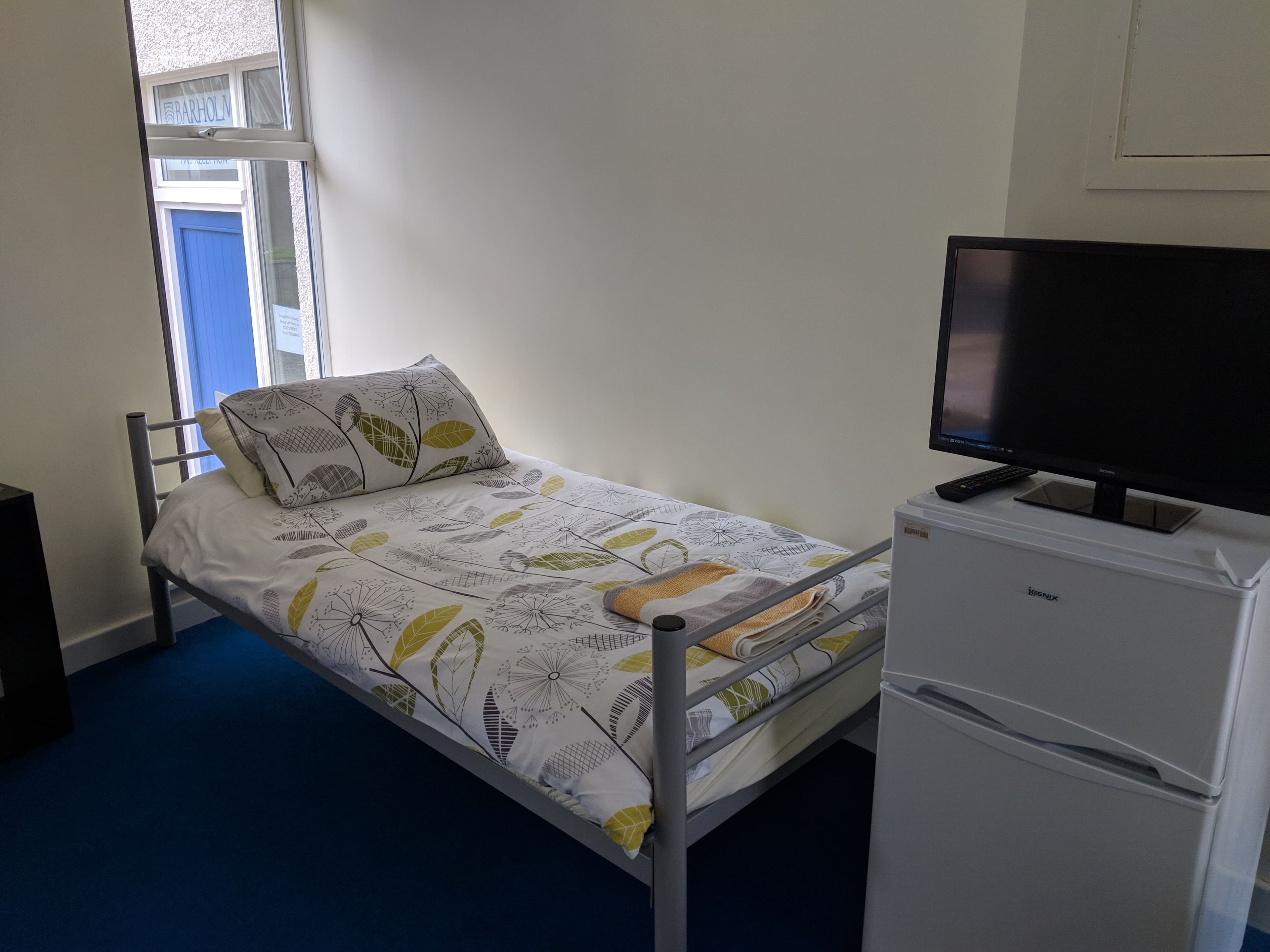 A single bed with a patterned bedspread, a television on a stand, and a white refrigerator.
