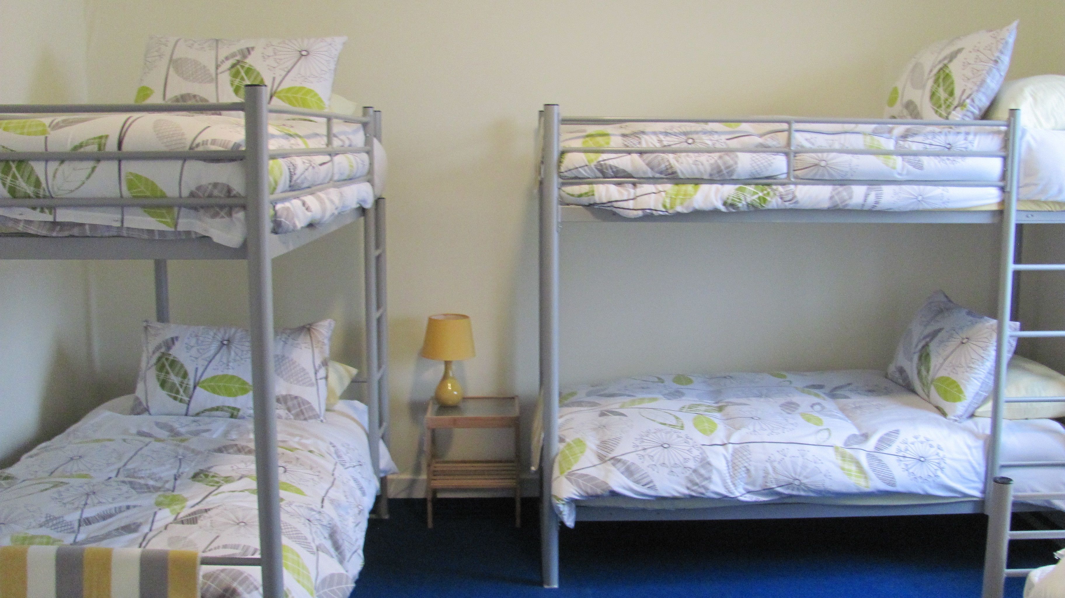Two bunk beds with leaf-patterned bedding, a small bedside table with a lamp, and a blue carpet.