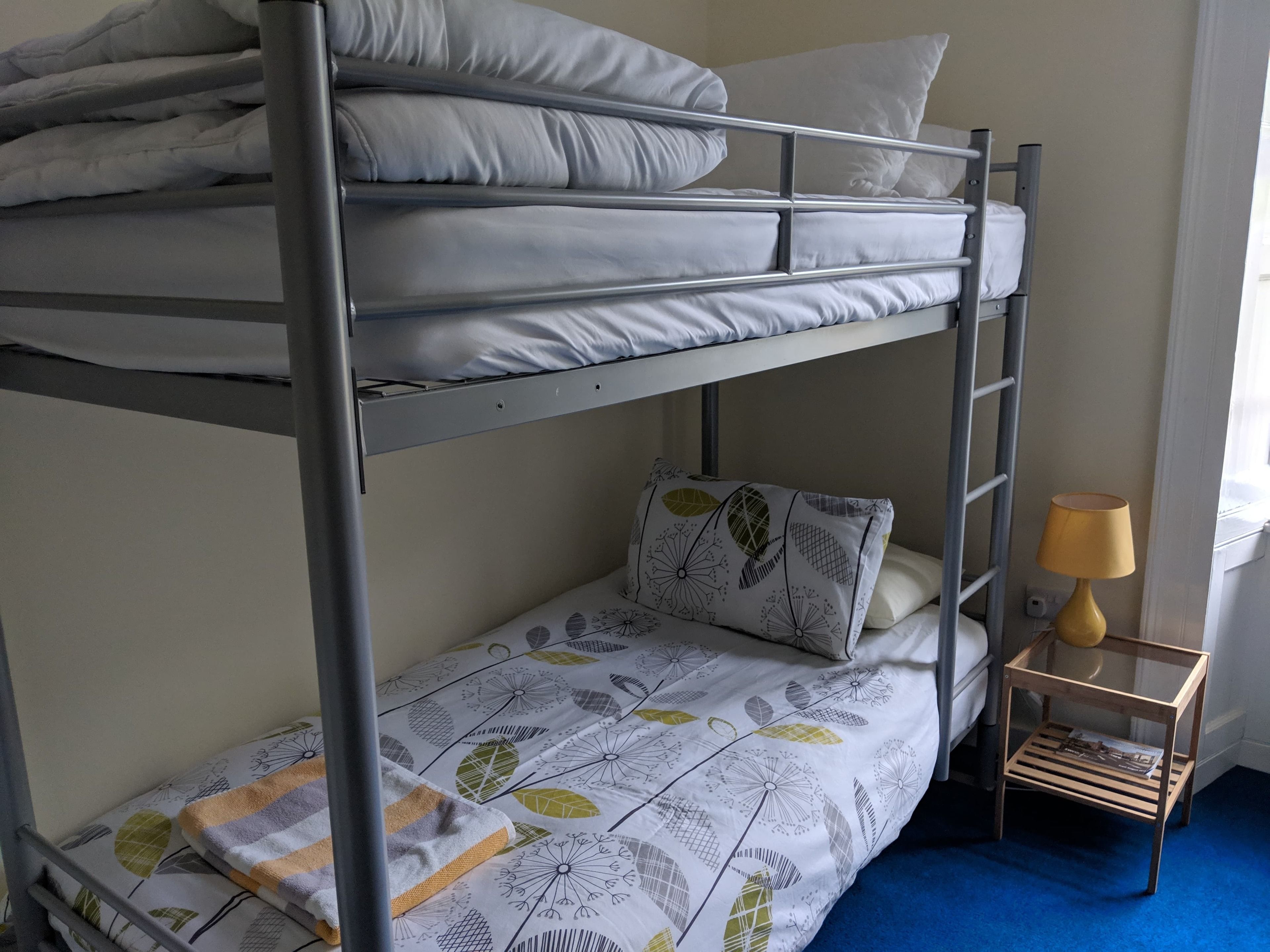 A bunk bed with a patterned bedspread and pillows, a small wooden nightstand with a lamp, and a blue carpet.