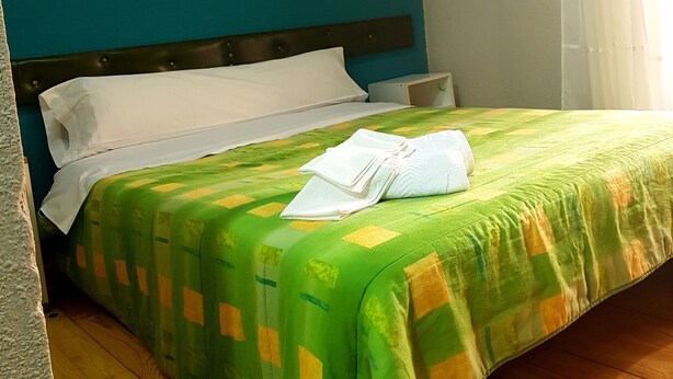 Un lit double recouvert d’une couvre-lit à motifs verts et jaunes, avec des oreillers blancs et une serviette blanche pliée posée sur le lit.