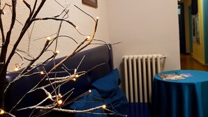 Un salon chaleureux avec un canapé bleu foncé, une petite table ronde sur laquelle sont posés des magazines et une branche décorative illuminée de guirlandes lumineuses.