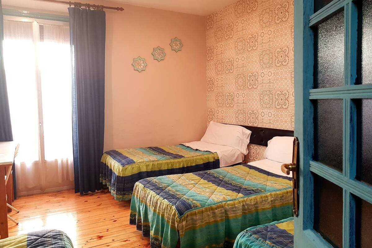 A room with two beds, a wooden floor, a window with curtains, and a patterned wall.