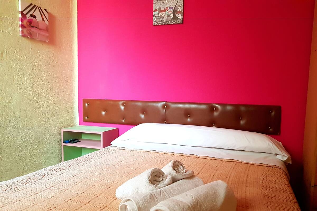 A hotel room with a pink wall, a brown headboard, a bed with white linens, and a green nightstand with a lamp.